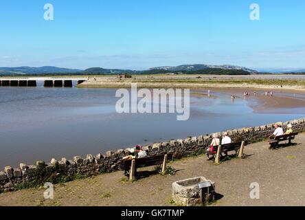 Menschen sitzen auf Arnside Strandpromenade Blick Flut im Mündungsgebiet des Flusses Kent auf Arnside Eisenbahnviadukt Arnside, Cumbria Stockfoto