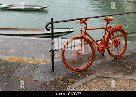 eine alte, ganz orange lackiert Fahrrad angebracht zu einem Metallgitter auf dem Bürgersteig in der Nähe eines Flusses Stockfoto