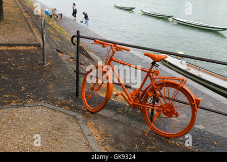 eine alte, ganz orange lackiert Fahrrad angebracht zu einem Metallgitter auf dem Bürgersteig in der Nähe eines Flusses Stockfoto