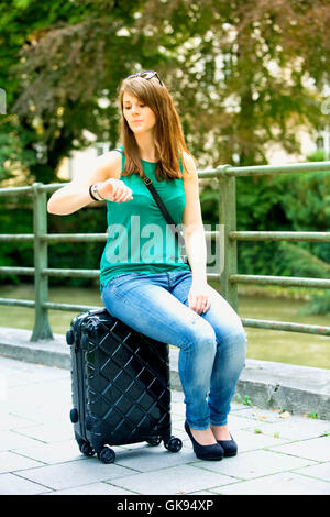 junge Brünette Frau sitzt auf dem Koffer und mit Blick auf ihre Uhr Stockfoto