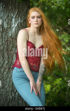 Schöne rote Kopf Frau in Röhrenjeans und rotes Top, fast vollen Länge gegen einen Baum gelehnt. Stockfoto