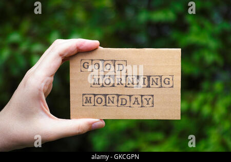 Frau Hand mit Pappkarte mit Worten guten Morgen Montag von schwarzen Alphabet Stempel gegenüber grünen Blumen Hintergrund gemacht. Stockfoto