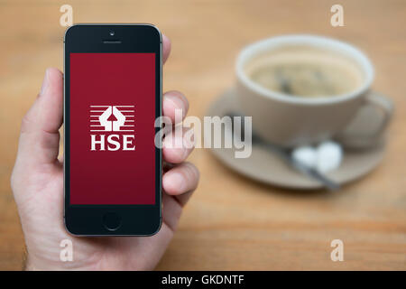 Ein Mann schaut auf seinem iPhone zeigt die HSE-Logo, während sie Sitzen mit einer Tasse Kaffee (nur redaktionelle Nutzung).