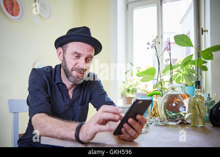 Hipster mit Bart und Hut Iphone sitzt auf einem Tisch in seiner Ktichen betrachten Stockfoto