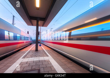 High-Speed Personenzüge auf Eisenbahn-Plattform in Bewegung in der Abenddämmerung. Verschwommene Zug. Bahnhof in der Nacht Stockfoto