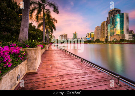 Moderne Wolkenkratzer, Blumen und Palmen entlang Lake Rajada bei Sonnenuntergang im Benjakiti Park in Bangkok, Thailand. Stockfoto