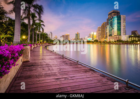 Moderne Wolkenkratzer, Blumen und Palmen entlang Lake Rajada bei Sonnenuntergang im Benjakiti Park in Bangkok, Thailand. Stockfoto