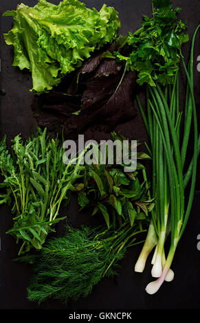 Verschiedene frische Bio-Kräuter (Kopfsalat, Rucola, Dill, Minze, rote Salat und Zwiebel) auf schwarzem Hintergrund im rustikalen Stil. Ansicht von oben Stockfoto