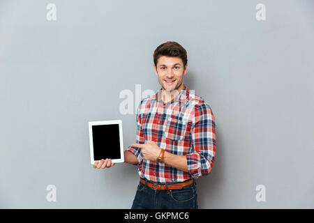 Fröhlichen jungen Mann im karierten Hemd, auf leeren Bildschirm Tablet über grauen Hintergrund zeigend Stockfoto