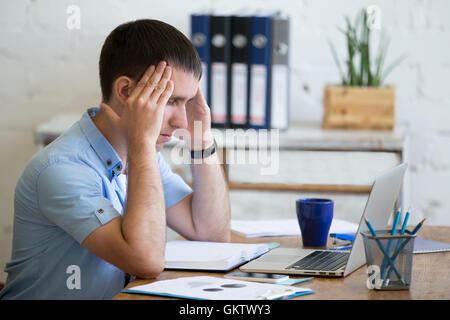 Jung betonte Geschäftsmann vor Laptop sitzen und seinen Kopf mit gequälten Ausdruck zu berühren. Business Mann Schmerzen Stockfoto