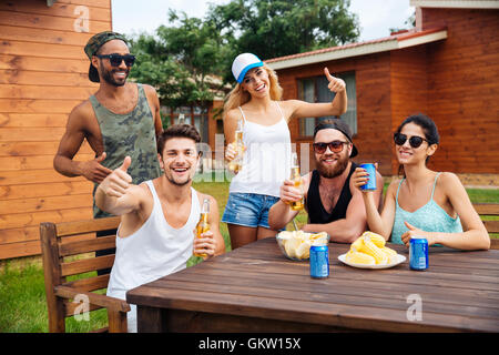 Gruppe von fröhlichen Jugendlichen Bier trinken und zeigt Daumen sich an den Tisch im freien Stockfoto