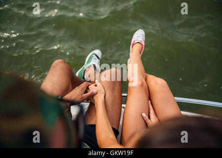 Draufsicht der Beine des jungen Paares in Turnschuhen sitzt nahe am Meer Stockfoto