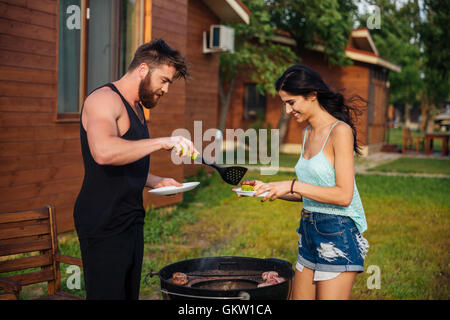 Glückliches junges Paar stehen und Kochen treffen auf Barbecue-Grill im freien Stockfoto