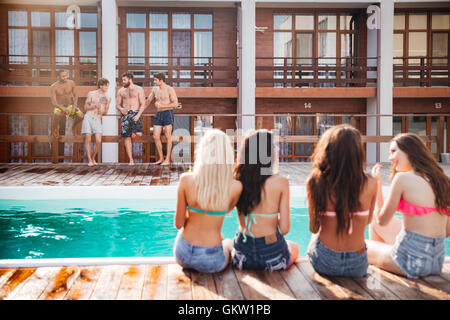 Rückansicht des attraktiven jungen Frauen sitzen in der Nähe von Schwimmbad und Blick auf attraktive Männer in kurzen Hosen Stockfoto
