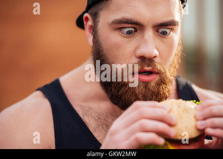 Nahaufnahme von erstaunt schockiert bärtiger junger Mann halten und mit Blick auf den Hamburger im freien Stockfoto