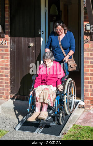 Eine 96 Jahre alte Dame lässt Ihr betreutes Wohnen in Ihrem Rollstuhl über eine Rampe mit ihrer Pflegeperson oder Assistent. Stockfoto