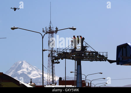 El Alto, Bolivien, 23. August 2016.Technicians warten, bis eine Drohne, die eine synthetische Lichtkabel zwischen den Masten einer neuen Seilbahn trägt / Gondelbahn, die befindet sich im Aufbau. Dies ist der erste Teil des Prozesses, das endgültige Stahlseil zu installieren, das Gondeln tragen wird. Seilbahn zwischen Rio Seco und La Ceja in El Alto gehört zu einer zweiten Phase der Seilbahn-Linien, die Teil eines ehrgeizigen Projekts um Verkehrsstaus zu entlasten. 3 Routen aus der ersten Phase sind bereits zwischen den Städten La Paz und El Alto in Betrieb. Mt Huayna Potosi ist im Hintergrund. Stockfoto