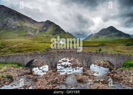 Eine alte Steinbrücke, unterstützt durch schroffe Berge und eine einsame Hütte unter einem dramatischen Stimmungsvoller Himmel Stockfoto