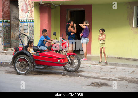 Kubanische Mütter stehen sprechen, während Ihre Kinder auf einer geparkten Motorrad und Seitenwagen in den Straßen von Centro Havanna Kuba spielen Stockfoto