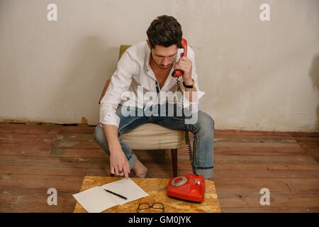 junger Mann sitzt auf dem alten Sessel und kommuniziert mit jemand. Es gibt Copybook, Stift, Gläser und Telefon auf der Palette Stockfoto