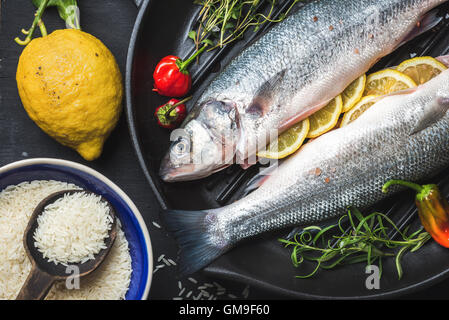 Rohe ungekochte Seebarsch mit Reis, Zitrone, Kräutern und Gewürzen auf schwarzes Grillen Eisen Pfanne auf dunklem Hintergrund Stockfoto