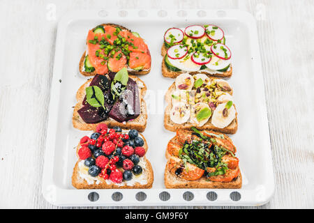 Süßes und herzhaftes Frühstück Toast Vielfalt. Sandwiches mit Obst, Gemüse, Eiern, geräucherter Lachs auf Backblech in hellem Holz Hintergrund weiß Stockfoto