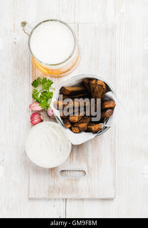 Bier-Snack-Set. Pint Pilsener in Becher und Schwarzbrot-Croûtons mit Knoblauch-Käse-Sahne-Sauce serviert frische Kräuter über weiß bemalten alten hölzernen Hintergrund Stockfoto