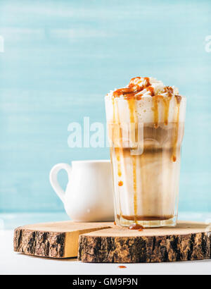 Latte Macchiato mit Schlagsahne Sahne-Karamell-Sauce im Longdrinkglas auf Holzbrett über blau gestrichenen Wand Hintergrund, Textfreiraum Stockfoto