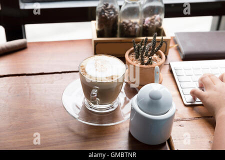 Hand tippen auf der Computertastatur mit heißen Kaffee Cappuccinotasse Stockfoto