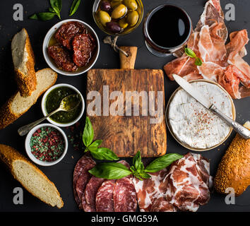 Wein snack Set mit leeren Holzbrett in Mitte. Glas Rot, Fleischauswahl, mediterranen Oliven, getrocknete Tomaten, Baguette Scheiben, Camembert Käse und Gewürze auf schwarzem Hintergrund, Stockfoto