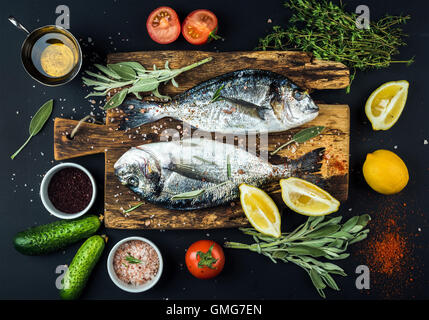 Ungekocht Dorado oder Dorade Fisch mit Zitrone, Kräutern, Öl, Gemüse und Gewürzen auf rustikalem Holzbrett auf schwarzem Hintergrund Stockfoto