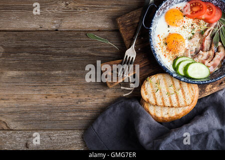 Frühstücks-Set. Pfanne mit Spiegelei mit Speck, frische Tomaten, Gurken, Salbei und Brot auf rustikalen Portion Brett über hölzerne Hintergrund Stockfoto