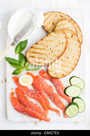Zutaten für gesunde Sandwich. Gegrillten Brotscheiben, Räucherlachs, Quark, Gurke und Basilikum auf weißen Holzbrett Stockfoto