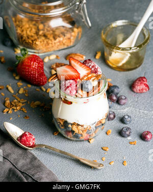 Joghurt-Hafer-Müsli mit frischen Beeren, Nüssen, Honig und Minze im Glas Glas auf grauem Beton strukturiertem Hintergrund Stockfoto