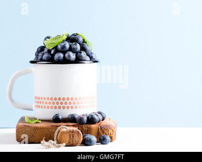 Frische reife Heidelbeeren im Land Stil Emaille Becher auf rustikalem Holzbrett über blau Pastell Hintergrund Stockfoto