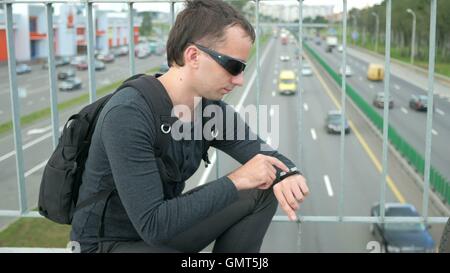 Outdoor Portrait moderner junger Mann mit smart-Uhr auf der Straße. Die in Gläser Rucksack sitzt auf der Brücke. Unten fahren Autos Stockfoto