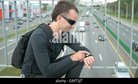 Outdoor Portrait moderner junger Mann mit smart-Uhr auf der Straße. Die in Gläser Rucksack sitzt auf der Brücke. Unten fahren Autos Stockfoto