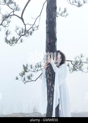 Junge Frau im weißen Kleid, eine Kiefer umarmt. Wiederherstellen der Verbindung mit der Natur. Stockfoto