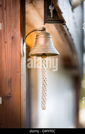 Eine kleine Schiffsglocke mit einem Seil verknotet Glocke wird auf einer Holzwand montiert. Eine Reihe von Tasten hängen oben.   Schiffsglocke. Stockfoto