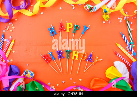 Happy Birthday Hintergrund mit verzierten Rahmen mit Partydekoration auf einem hellen orange Holztisch mit alles Gute zum Geburtstag Kerze Stockfoto