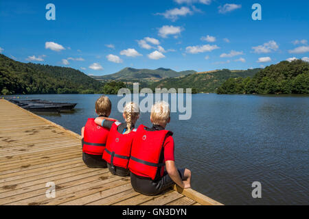 Drei Kinder in Schwimmwesten sitzen an einem Dock und blicken an einem sonnigen Tag auf den ruhigen See Chambon. Puy de Dome, Auvergne, Frankreich. Europa Stockfoto