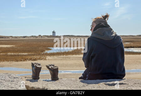Eine junge Frau sitzt auf einem Felsen-Steg mit ihren Stiefeln neben ihr und ein Leuchtturm in der Ferne.