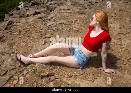 Schöne Frau mit langen roten Haaren, in Jeans-Shorts und rote Hemd, sitzen auf den Felsen des Berggipfel im Sommer. Stockfoto