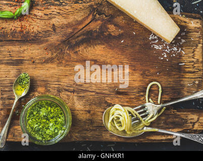 Italienische Küche kochen Hintergrund. Glas mit Pesto-Sauce, Löffel und Gabel mit gekochten Spaghetti, frischem Basilikum und Parmesan Stockfoto