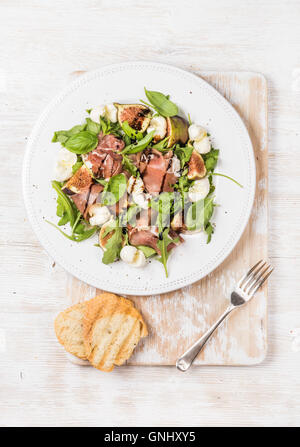 Schinken, Rucola, Basilikum, Feigen und Mozzarella-Salat serviert mit Brotscheiben auf rustikalen Brett über weißen hölzernen Hintergrund, Stockfoto