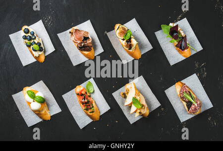 Italienische Crostini mit verschiedenen Toppings auf Weißes Backpapier über schwarze Sperrholz Hintergrund, Ansicht von oben Stockfoto