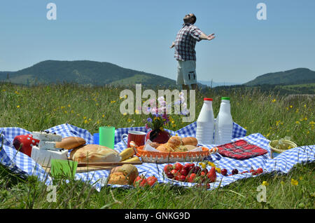 Mann sein Gesicht zur Sonne drehen und verbreitet seine Arme hinter eine Picknick-Set-up auf Wiese mit Hügeln im Hintergrund Stockfoto