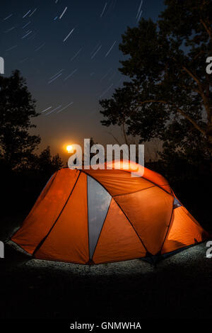 Langzeitbelichtung von einem Zelt in der Nacht mit zunehmender Mond am Horizont und Sterne Wanderwege in den Nachthimmel. Stockfoto