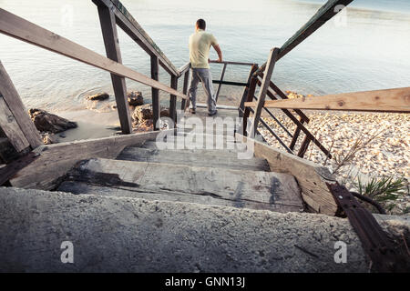 Junger Mann steht auf alte Holztreppe hinunter zur Küste Meeres Stockfoto
