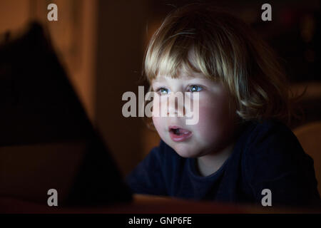 Blondine zwei Jahre alten baby-blauen Pullover, Haus in der Nacht Lesung sitzen und beobachten digital-Tablette, Gesicht beleuchtet von th Stockfoto
